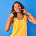 A brown woman points to her beautiful smile while wearing a yellow tank top against a blue wall