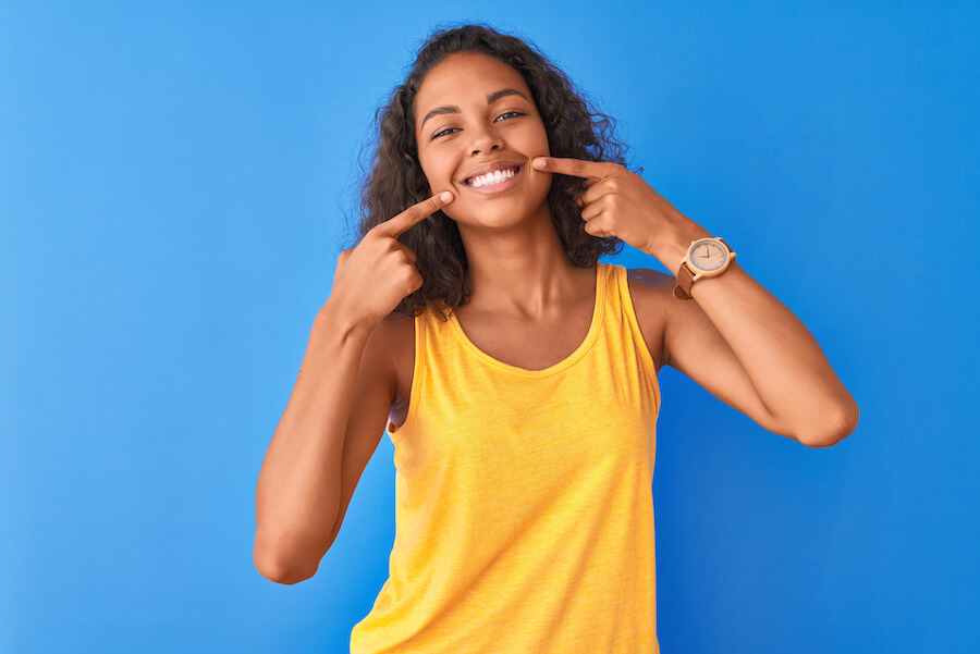 A brown woman points to her beautiful smile while wearing a yellow tank top against a blue wall