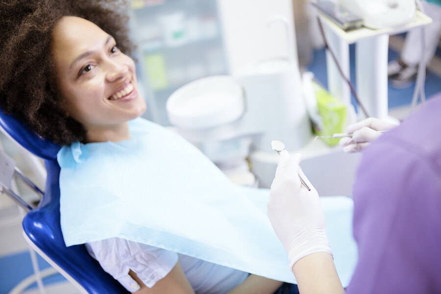 Black woman sits in the dental chair and smiles at her dental hygienist before her routine teeth cleaning
