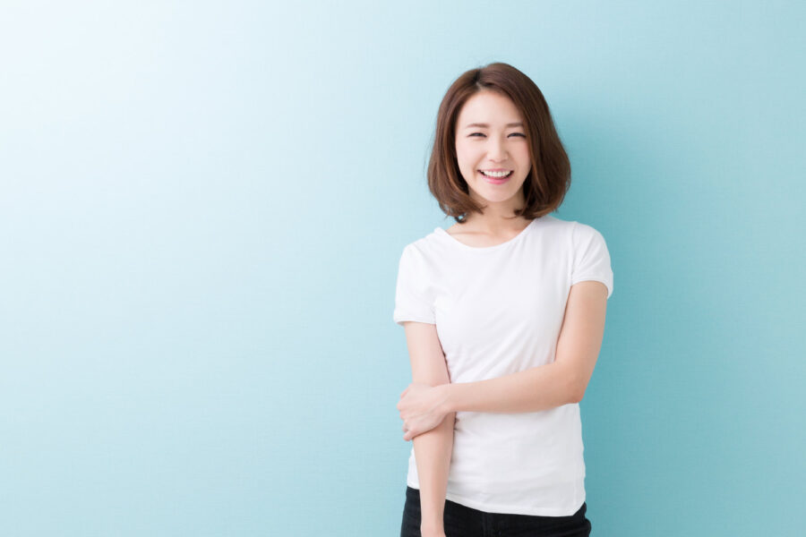 Asian woman with veneers in a white t-shirt smiles while standing against a blue wall