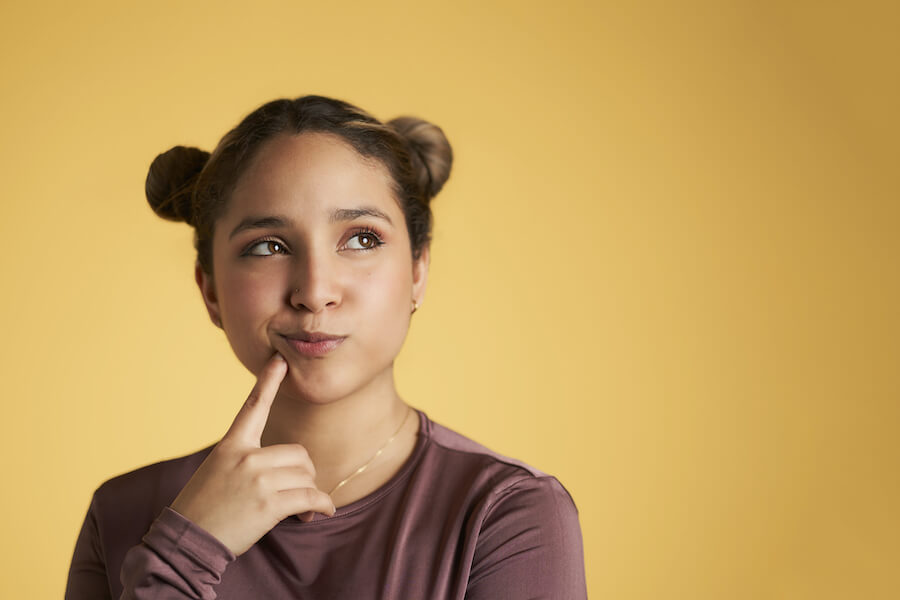 Young girl with space buns hair looks questioning with her index finger resting on the corner of her mouth, common causes of tooth stains, teeth stains